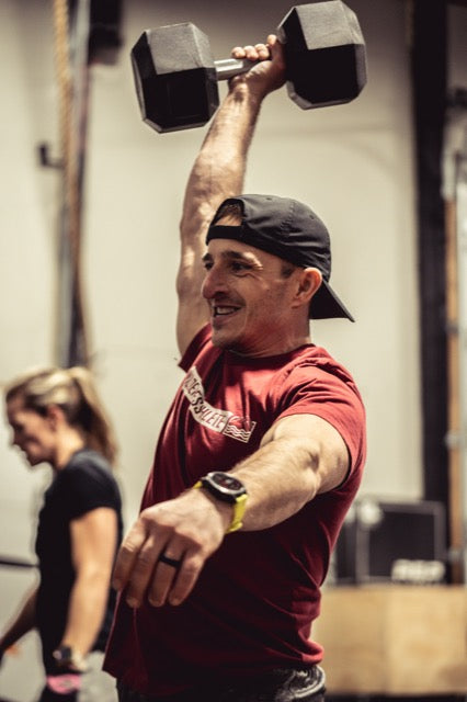 man in red shirt lifting dumbbell overhead with woman in the background